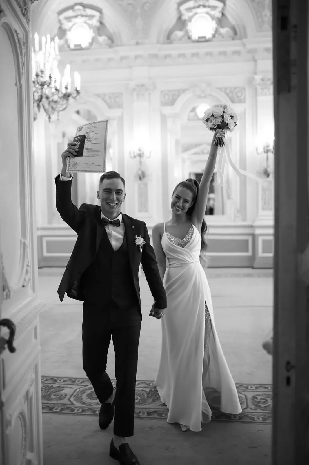 Les mariés avancent bras levés dans une grande salle aux moulures blanches : lui brandit un document, elle un bouquet blanc. Photo en noir et blanc.